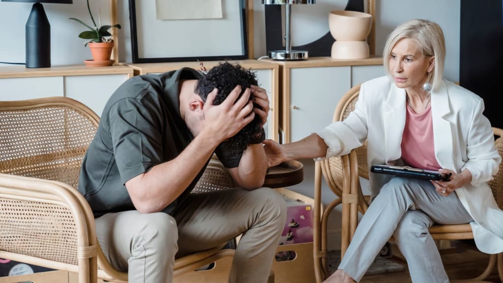 A person sitting and holding their head in distress while another person sits beside them, offering support by placing a hand on their shoulder in a cozy room.